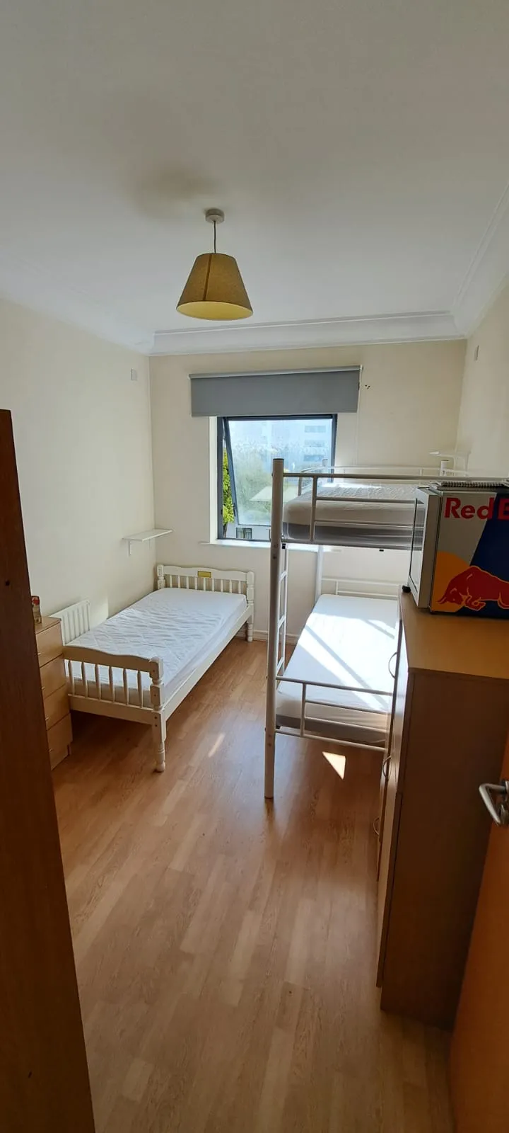 Bedroom with a single bed and a bunk bed, window with a roller blind, wooden floor, dresser and ceiling light.