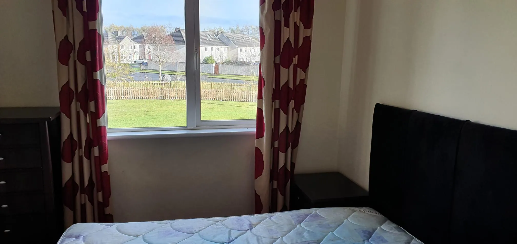Bedroom with a single bed, burgundy patterned curtains, and a large window overlooking a garden, fence, and neighboring houses.