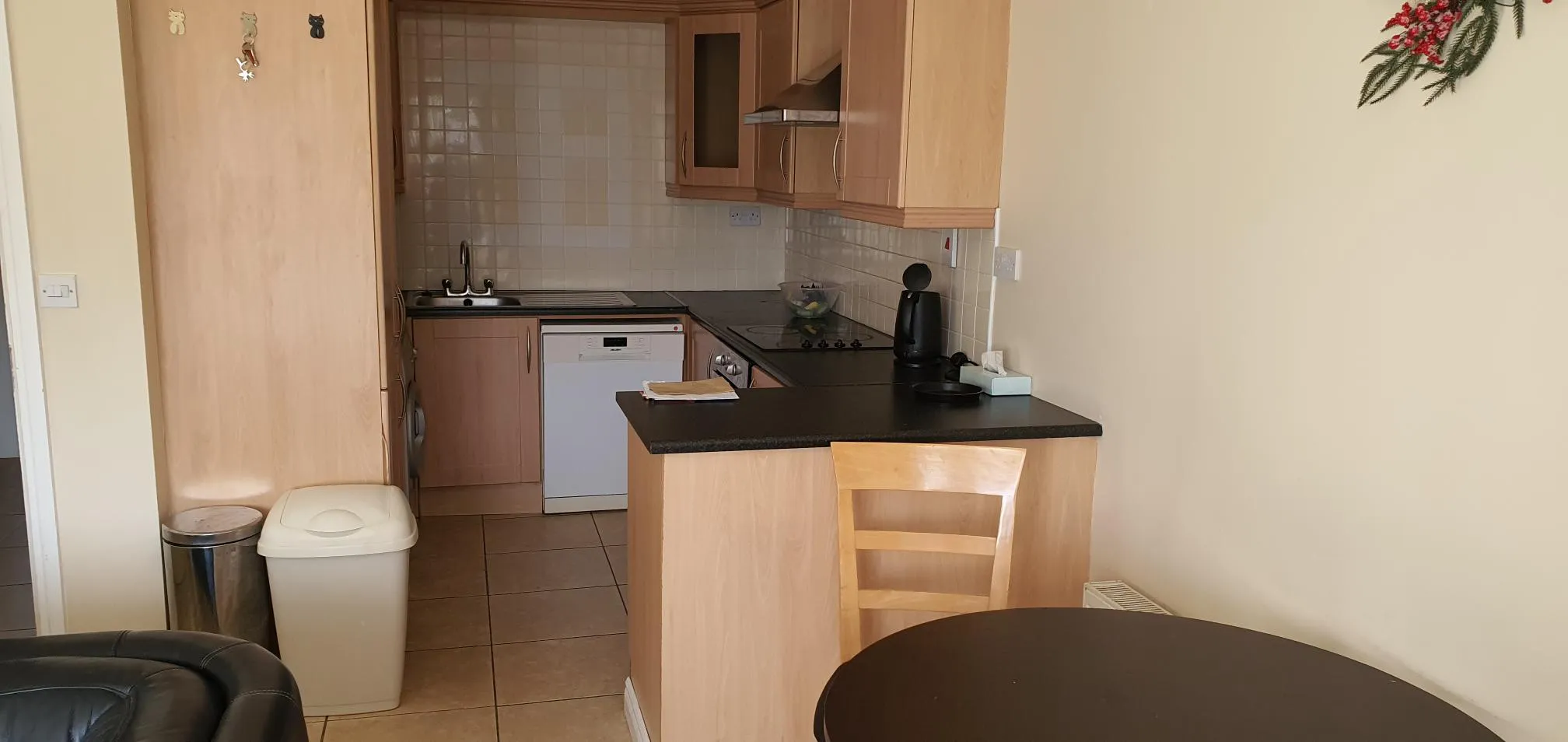 Kitchen with wooden cabinets, black countertop, gas hob, white dishwasher, sink, and breakfast bar with black top. Beige walls and tiled floor.