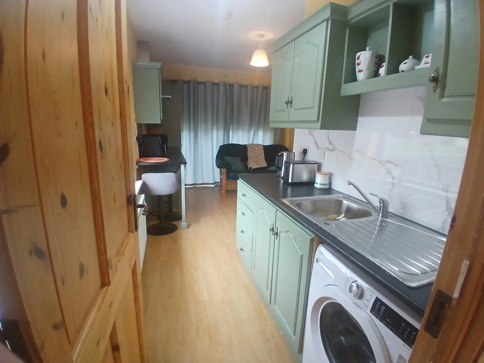 Narrow kitchen with green cabinets, countertop sink, washing machine, toaster and a small seating area visible through the doorway