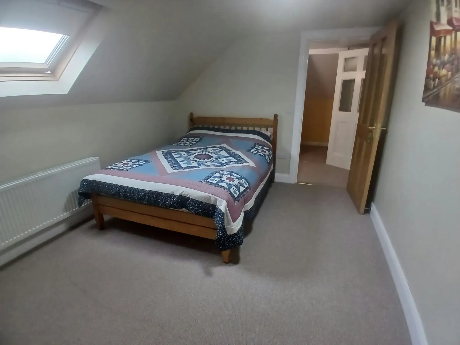 Bedroom with a single/double wooden bed with patterned quilt, skylight window, radiator along the left wall, and an open door to a hallway.