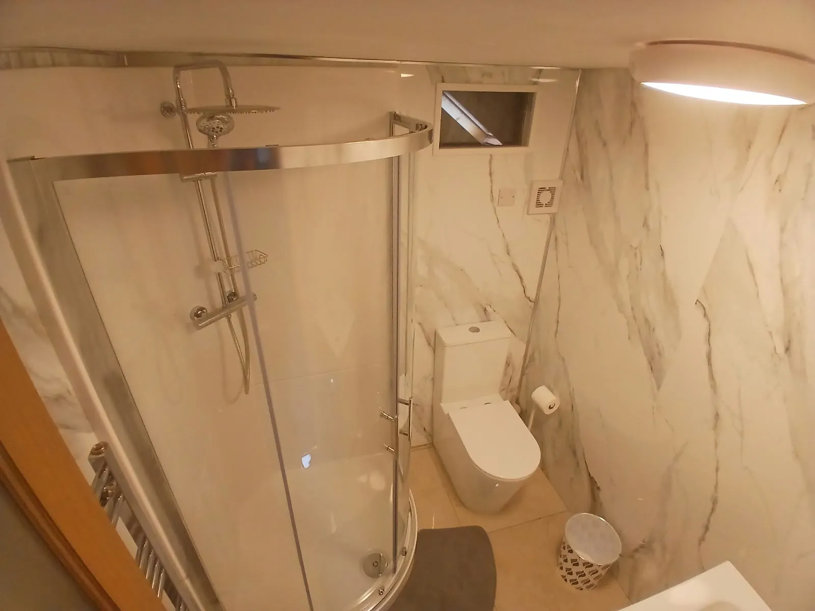 Bathroom with a curved glass shower enclosure and wall-mounted showerhead, a toilet, marble-patterned walls, ceiling light, and a small waste bin.
