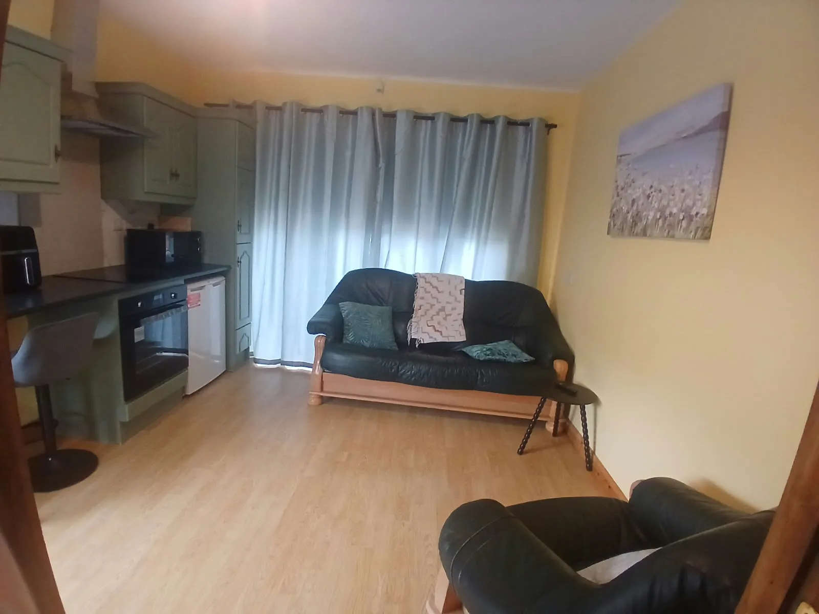 Living room with a dark leather two-seater sofa, armchair, side table, large curtained window and a small open kitchenette along the left wall.