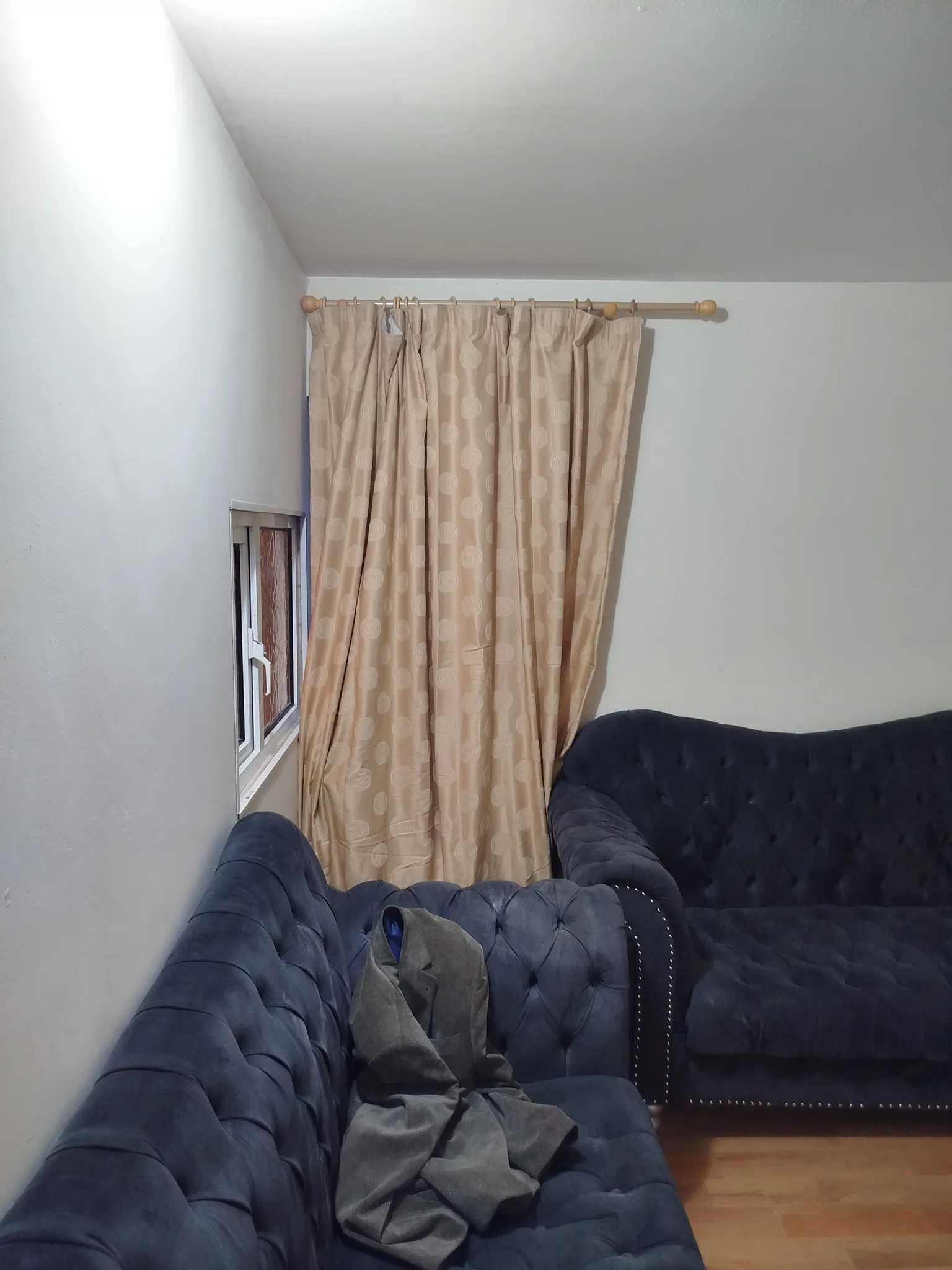 Living room corner with beige curtains covering a small window, a wooden curtain rod, and two dark blue tufted sofas; a jacket is draped on one sofa.