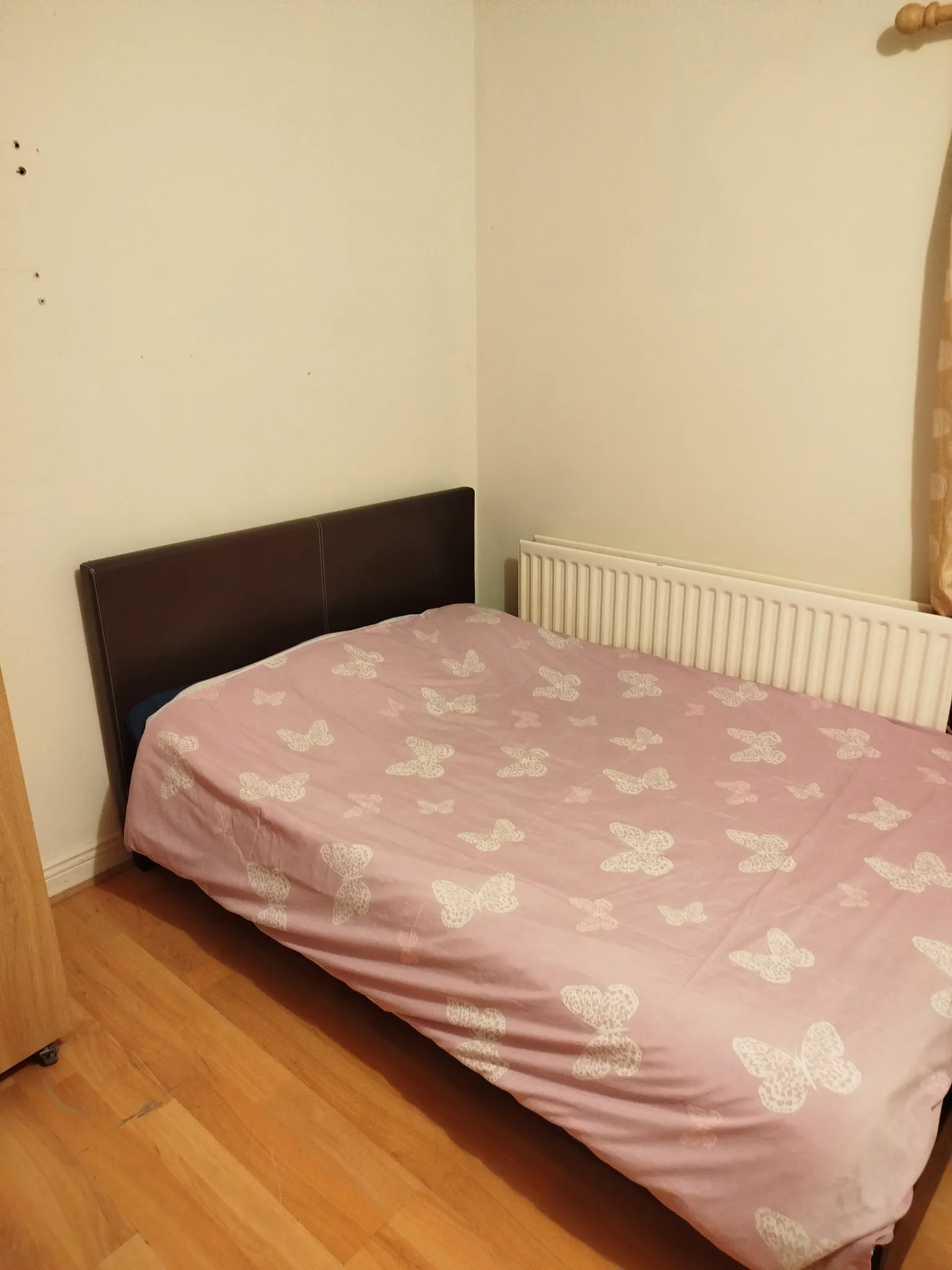 Bedroom corner with a double bed covered by a pink butterfly duvet, a dark headboard, a white radiator and a curtain at the right edge.