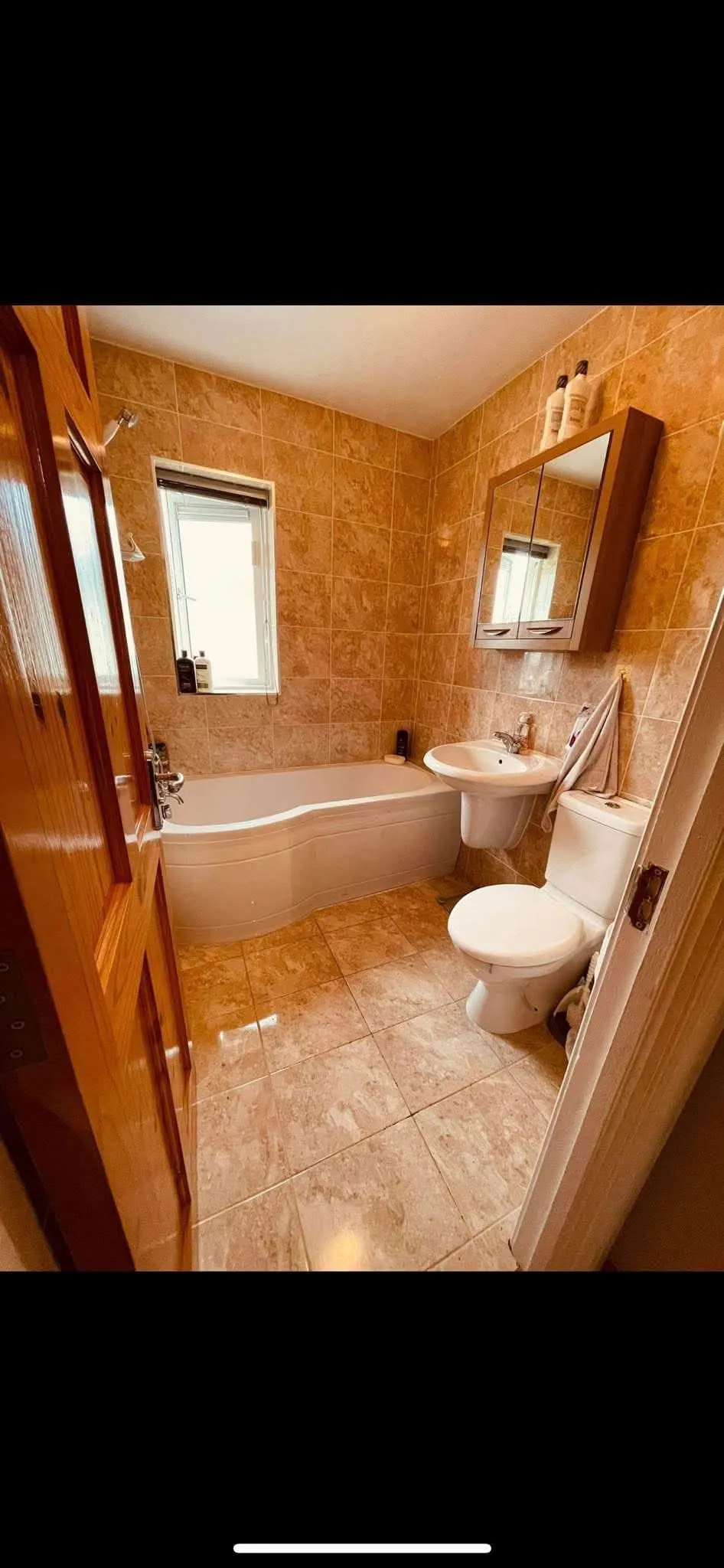 Bathroom with a bathtub along the left wall, wall-mounted sink and mirror cabinet, and a toilet on the right; tiled walls and floor and a small window above the tub.